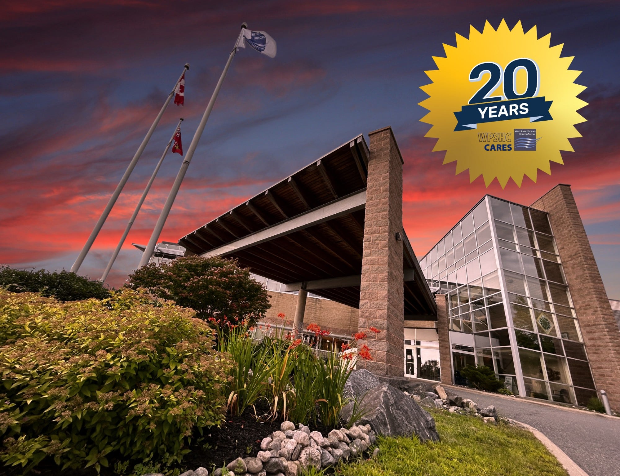 West Parry Sound Health Centre’s main entrance looms large in front of the viewer, as flags wave in the breeze. In the background a vivid sunset fills the sky. Superimposed on the image in the corner is a badge celebrating 20 years and the WPSHC Cares logo.