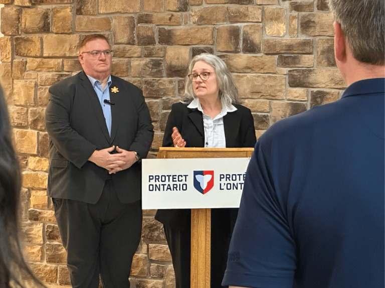 A bespectacled woman in a suit stands behind a podium that says "Protect Ontario," while gesturing and speaking. A bespectacled man in a suit stands behind her, to her left, listening.